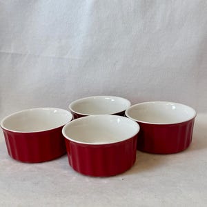 May include: A set of four round ramekins featuring a glossy deep red exterior and a smooth white interior. Each ceramic dish has a classic fluted side design with vertical ridges, providing a consistent look for these small, open-top baking vessels.