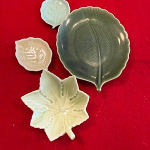 May include: Four ceramic leaf-shaped dishes in varying shades of green and off-white. The largest dish is a deep green with a prominent central vein. The other dishes are smaller, with detailed leaf vein patterns. The dishes are arranged on a red surface.