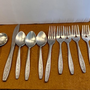 May include: A collection of stainless steel silverware, including spoons, forks, and a knife, arranged on a brown surface. The silverware has a floral design on the handles. The set includes a variety of utensils for a complete dining experience.