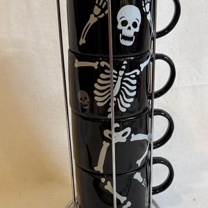 May include: A set of four black ceramic mugs stacked in a metal rack. Each mug features a white skeleton design. The mugs are cylindrical with a handle and are designed for beverages. The set is ideal for Halloween or spooky-themed decor.