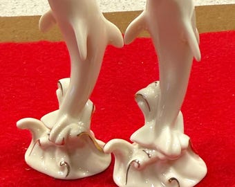 May include: Two ivory-colored porcelain dolphin figurines. The dolphins are leaping, with open mouths and detailed fins. They are mounted on a base with gold accents, set against a red background. These decorative sculptures are ideal for home decor.
