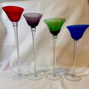 May include: Four colorful glass candle holders. The candle holders have a clear, slender stem and a flared bowl in red, purple, green, and blue. The candle holders are arranged in a row.