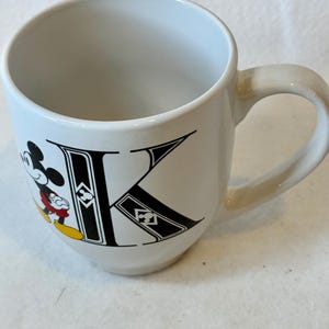 May include: White ceramic mug with a black "K" and a Mickey Mouse illustration. Mickey is wearing red shorts, yellow shoes, and black gloves. The mug has a curved handle and a smooth, glossy finish.