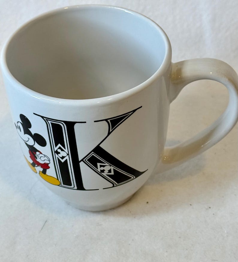 May include: White ceramic mug with a black "K" and a Mickey Mouse illustration. Mickey is wearing red shorts, yellow shoes, and black gloves. The mug has a curved handle and a smooth, glossy finish.