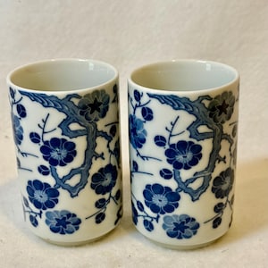 May include: Two cylindrical ceramic cups featuring a white base with a repeating blue floral pattern of plum blossoms on gnarled branches. The cups have a smooth, straight-sided design with an open top and a solid, flat base for stability.