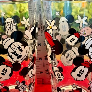 Puede incluir: Dos vasos transparentes decorados con im&aacute;genes de Mickey y Minnie Mouse, junto con flores. Los vasos tienen un patr&oacute;n repetido de los personajes de dibujos animados en negro, blanco y rojo. Una regla se coloca entre los vasos.