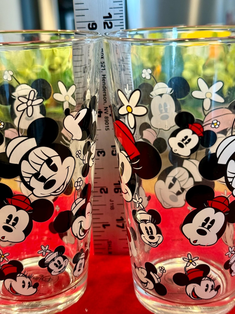 Puede incluir: Dos vasos transparentes decorados con im&aacute;genes de Mickey y Minnie Mouse, junto con flores. Los vasos tienen un patr&oacute;n repetido de los personajes de dibujos animados en negro, blanco y rojo. Una regla se coloca entre los vasos.