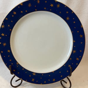 May include: A round ceramic plate featuring a plain white center and a wide, deep blue border decorated with various gold-colored star shapes of different sizes. The outer and inner edges of the blue rim are finished with a thin gold-colored line.