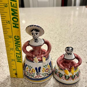May include: Two ceramic figurines of women wearing traditional dresses with floral patterns. The larger figurine is wearing a hat and has a red dress with blue and yellow floral accents. The smaller figurine is wearing a red dress with green and yellow floral accents.