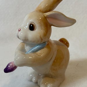 May include: A ceramic bunny figurine with tan and white fur, holding a purple paintbrush and a blue scarf. The bunny has large brown eyes and a pink nose, with its ears perked up. The figurine is likely a decorative item.