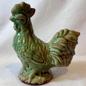 May include: A ceramic rooster figurine in shades of green and brown. The rooster has a detailed, textured surface with a crackle glaze effect. The figurine is standing on a small base.