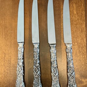 May include: Four silver-toned dinner knives with smooth, polished metal blades and ornate, textured handles featuring a detailed relief pattern of intertwined grapevines, leaves, and clusters of grapes extending along the entire length of the handle.