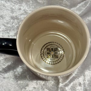 May include: Cream-colored ceramic mug with a black handle. The interior features a decorative gold and black design at the bottom. The mug has a glossy finish and a simple, elegant design, suitable for hot beverages.