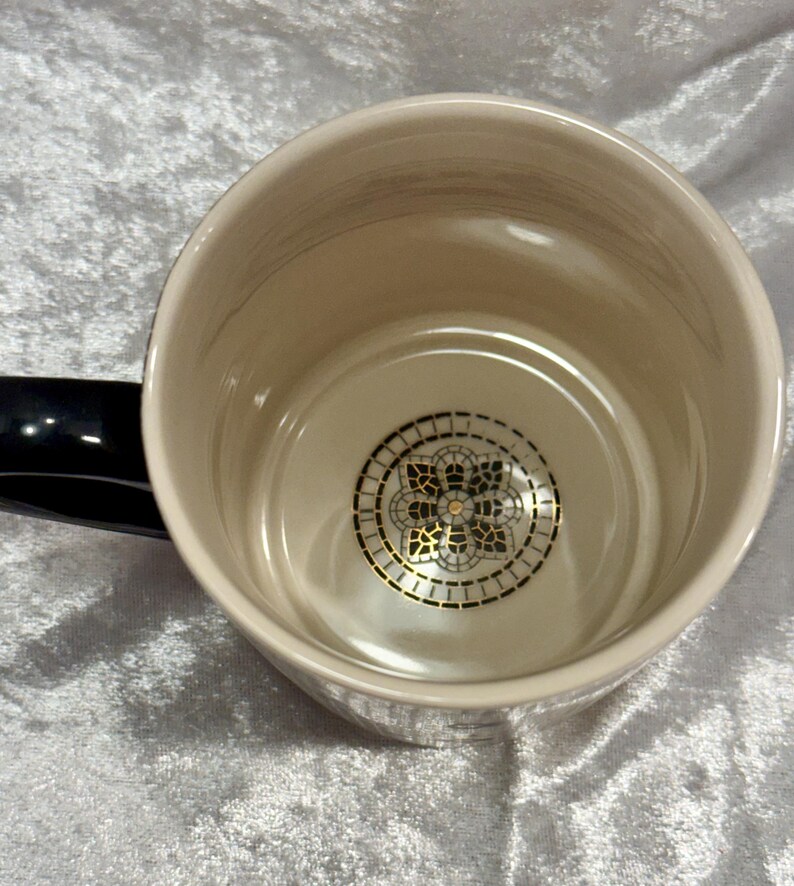 May include: Cream-colored ceramic mug with a black handle. The interior features a decorative gold and black design at the bottom. The mug has a glossy finish and a simple, elegant design, suitable for hot beverages.