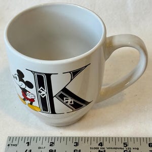 May include: White ceramic mug featuring Mickey Mouse and the letter "K" in black. Mickey is in his classic pose with red shorts and yellow shoes. The mug has a curved handle.