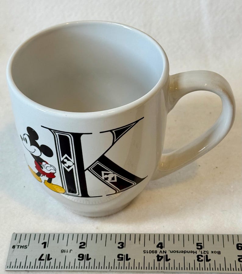 May include: White ceramic mug featuring Mickey Mouse and the letter "K" in black. Mickey is in his classic pose with red shorts and yellow shoes. The mug has a curved handle.