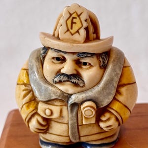 May include: A detailed figurine of a firefighter in a tan uniform with a tan helmet. The firefighter has a dark mustache and a stern expression. The helmet has a badge with the letter "F".