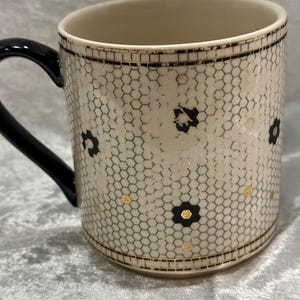 May include: Cream-colored ceramic mug with a black handle. The mug features a honeycomb pattern with black floral accents and gold details. The rim and base are trimmed with gold and black lines.
