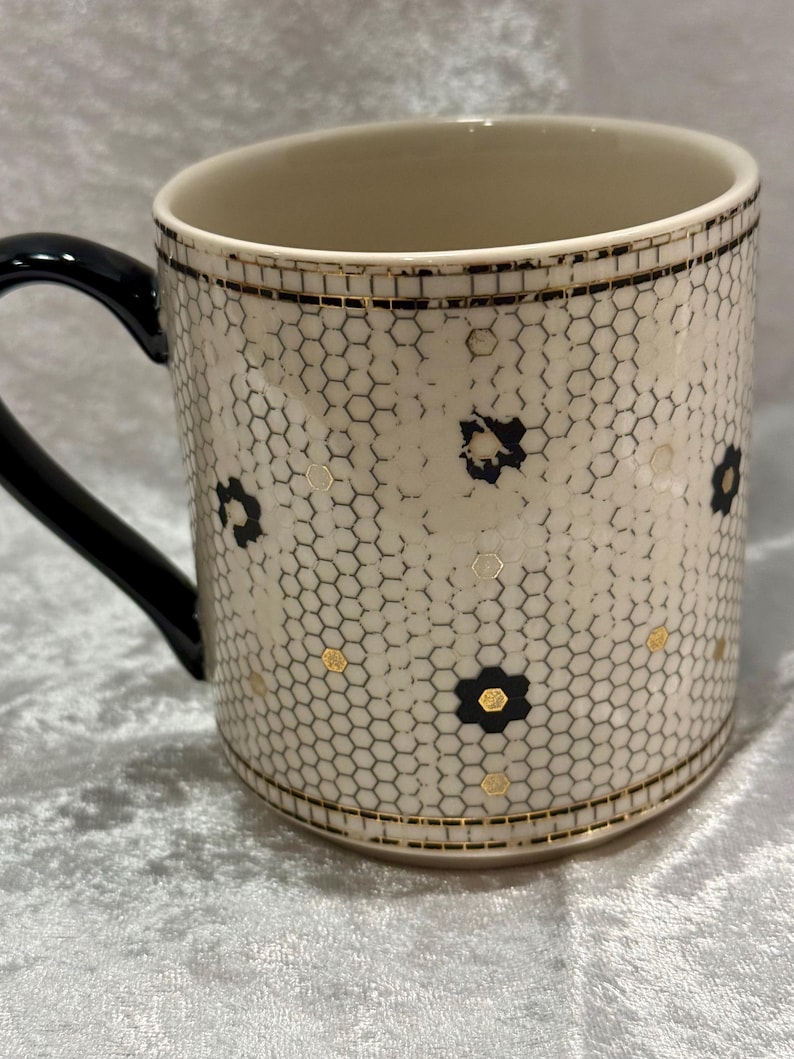May include: Cream-colored ceramic mug with a black handle. The mug features a honeycomb pattern with black floral accents and gold details. The rim and base are trimmed with gold and black lines.