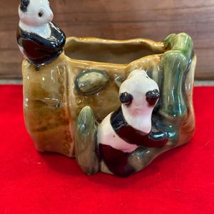 May include: A ceramic planter featuring two panda bear figurines. The planter is a rectangular shape with a brown exterior and green bamboo accents. One panda sits on the edge, while the other clings to the side. The pandas have black and white markings.