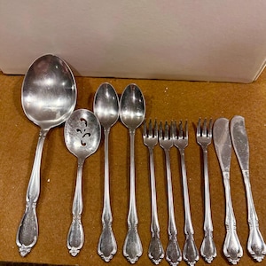 May include: A collection of vintage silverware, including spoons, forks, and knives, arranged on a brown surface. The silverware has ornate handles and a shiny, reflective surface. The set includes a serving spoon and a slotted spoon.
