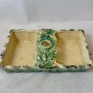 May include: A rectangular ceramic divided dish with a cream-colored interior and a green leaf-patterned center handle featuring a circular opening. The exterior edges are scalloped and finished with a dark, speckled, distressed-style trim.