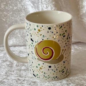 May include: A set of three ceramic mugs with a speckled, matte finish. The mugs are in shades of cream, light gray, and dark gray. Each mug has a simple, minimalist design, perfect for coffee or tea.