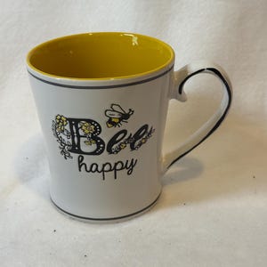 May include: A white ceramic mug with a yellow interior, featuring the text "Bee happy" in black lettering. The design includes a small bee illustration and floral accents. A ruler at the bottom shows the mug is approximately 4.5 inches tall.