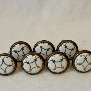May include: A set of seven round cabinet knobs featuring a white ceramic center with a gold-toned metallic lattice pattern. Each knob is encased in a circular antique brass-colored metal frame with a smooth, polished finish on the front surface.