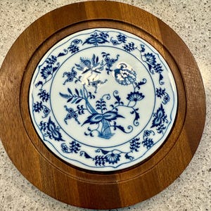 May include: A round wooden trivet with a white ceramic center. The ceramic has a blue floral pattern. The trivet is designed to protect surfaces from hot dishes. The wooden frame is a medium brown color.