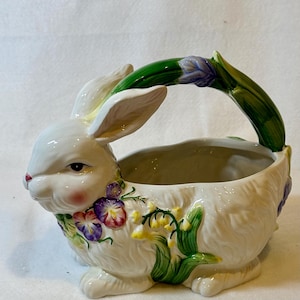 May include: A white ceramic bunny-shaped basket with a green handle and floral accents. The bunny has pink cheeks and is decorated with purple, red, and yellow flowers. The basket is designed for decorative use.