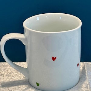 May include: White ceramic mug with a curved handle. The mug features a glossy finish and is decorated with small red and green heart-shaped designs. The mug is set against a blue background and a white surface.