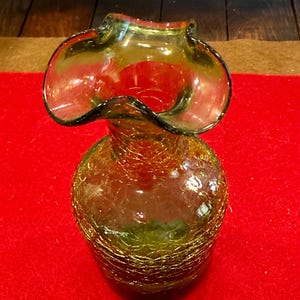 May include: A vintage, olive-green glass vase with a ruffled rim. The vase has a crackle finish and a round body. The rim is a darker green, and the vase sits on a red surface. The vase is likely used for decorative purposes.