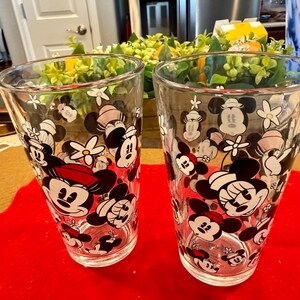 Puede incluir: Dos vasos transparentes con un patr&oacute;n repetido de personajes de Mickey y Minnie Mouse, junto con flores blancas. Mickey lleva un sombrero rojo y Minnie un lazo rosa. Los vasos est&aacute;n sobre una superficie roja.