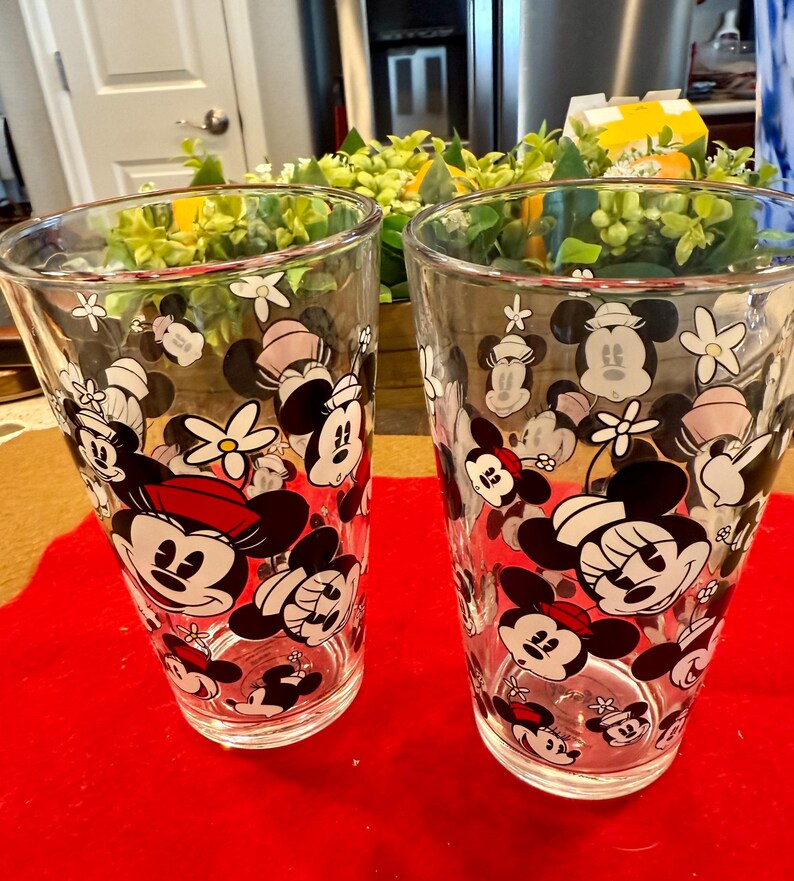 Puede incluir: Dos vasos transparentes con un patr&oacute;n repetido de personajes de Mickey y Minnie Mouse, junto con flores blancas. Mickey lleva un sombrero rojo y Minnie un lazo rosa. Los vasos est&aacute;n sobre una superficie roja.