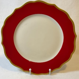 May include: A decorative plate with a cream-colored center and a vibrant red border. The rim has a scalloped edge with a gold trim. The plate is displayed on a white stand.