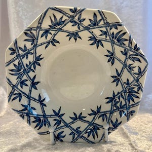 May include: An octagonal white plate with a blue bamboo pattern. The bamboo design features interconnected stalks and leaves, creating a lattice-like border around the plate's edge. The plate is displayed on a white stand.