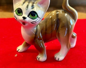 Puede incluir: Una figura de cerámica de un gato con expresión de sorpresa. El gato tiene ojos verdes, pecho blanco y rayas marrones y beige. La figura está sobre una superficie roja.
