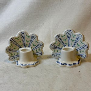 May include: Two white ceramic candle holders with a peacock feather design. The feather design features blue and green accents. Each holder has a cylindrical base and a scalloped edge.