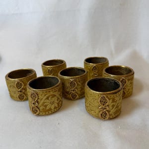 May include: A set of eight gold-colored napkin rings. Each ring features a detailed floral pattern and an ornate, decorative accent. The rings are cylindrical and appear to be made of a woven material.