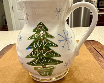 May include: White ceramic pitcher with a gold rim and handle. The pitcher is decorated with a green Christmas tree and blue snowflakes. The base of the pitcher has a gold trim.