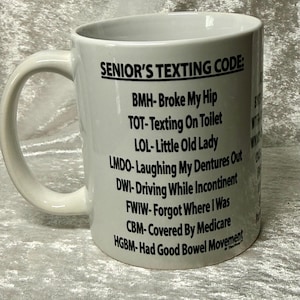 May include: White ceramic mug with a curved handle. The mug features the text "SENIOR'S TEXTING CODE:" followed by a list of acronyms and their meanings, including "BMH- Broke My Hip" and "TOT- Texting On Toilet."