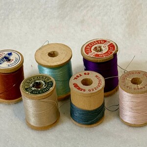 May include: Six vintage wooden spools of sewing thread in various colors including rust, light blue, beige, dark purple, dark teal, and pale pink. Each spool features a circular paper label on top with brand text like Clark's and J.P. Coats.