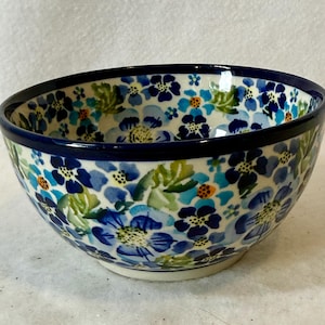 May include: A ceramic bowl with a dark blue rim and a floral pattern. The bowl features a white base with blue, green, and orange flowers. The flowers are in various shades of blue, with green leaves. The bowl is likely for serving food.