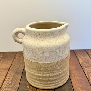May include: A ceramic pitcher with a handle and spout. The top half is speckled off-white, and the bottom half is a textured tan. The pitcher sits on a wooden surface.