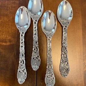 May include: Four silver-toned metal spoons featuring intricate, raised relief designs of grape clusters, vines, and leaves along the handles. The bowls of the spoons are smooth, polished, and reflective, while the handles exhibit a textured, ornate pattern.