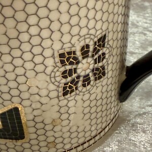 May include: Cream-colored ceramic mug with a black honeycomb pattern and a black handle. The mug features a decorative floral design with black and gold accents. The bottom of the mug has a geometric border.