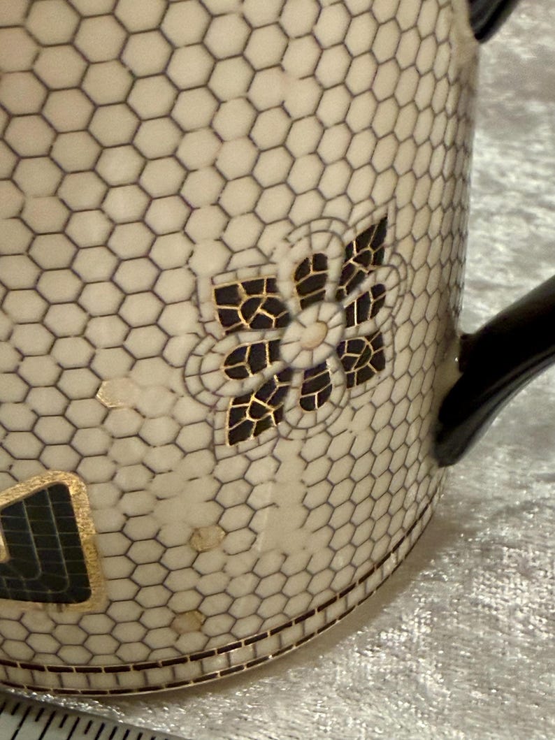May include: Cream-colored ceramic mug with a black honeycomb pattern and a black handle. The mug features a decorative floral design with black and gold accents. The bottom of the mug has a geometric border.
