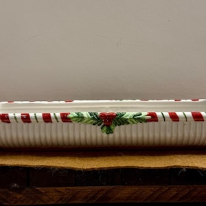 May include: A rectangular ceramic serving dish with a white base and red and green accents. Santa Claus and a reindeer figurine flank the dish. The dish has a candy cane stripe design and a holly and berry motif.