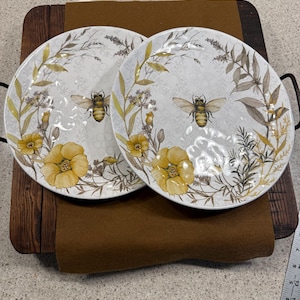May include: Two white ceramic plates with a bee and floral design. The plates feature yellow flowers, green leaves, and a bee in the center. The plates are on a brown surface.